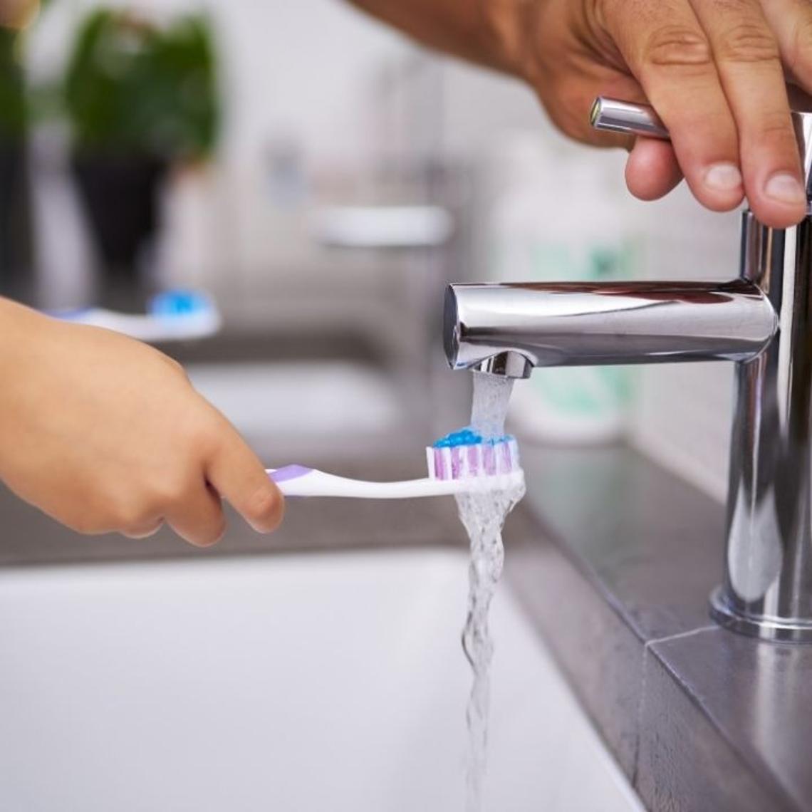 Parent turning on bathroom tap for child holding toothbrush under the tap